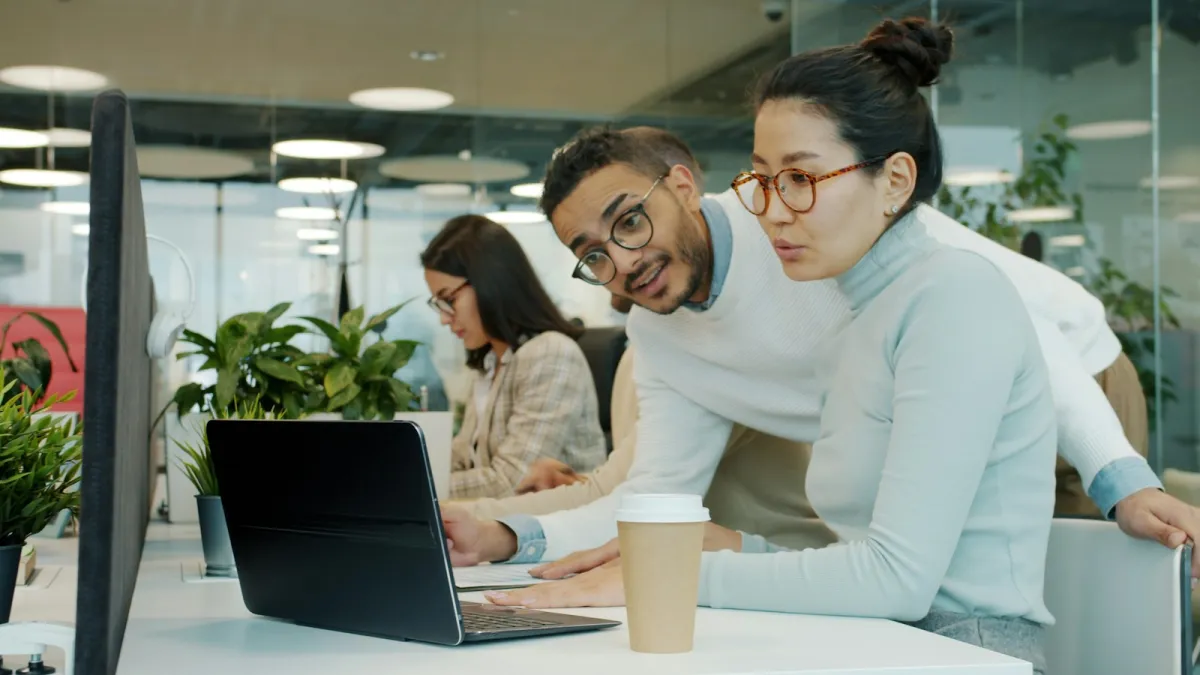 Two colleagues at a desk, one helping the other check something on a laptop